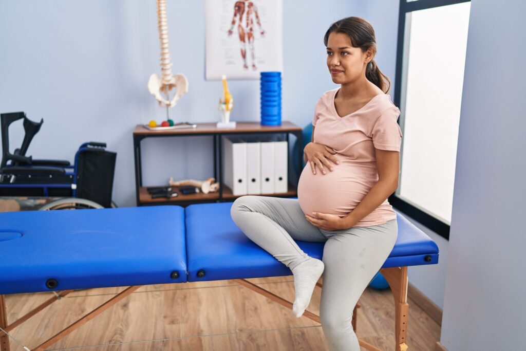 Pregnant woman sitting on a blue treatment table in a clinical setting with anatomical models and medical equipment visible in the background.