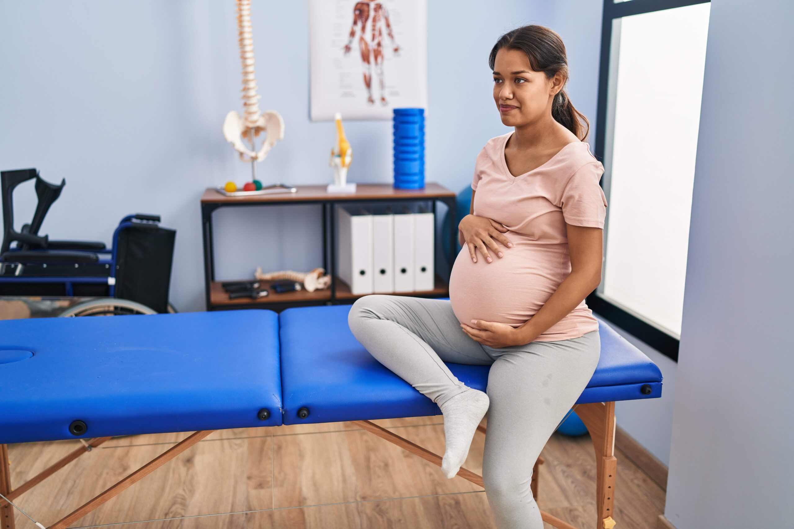 Pregnant woman sitting on a blue treatment table in a clinical setting with anatomical models and medical equipment visible in the background.