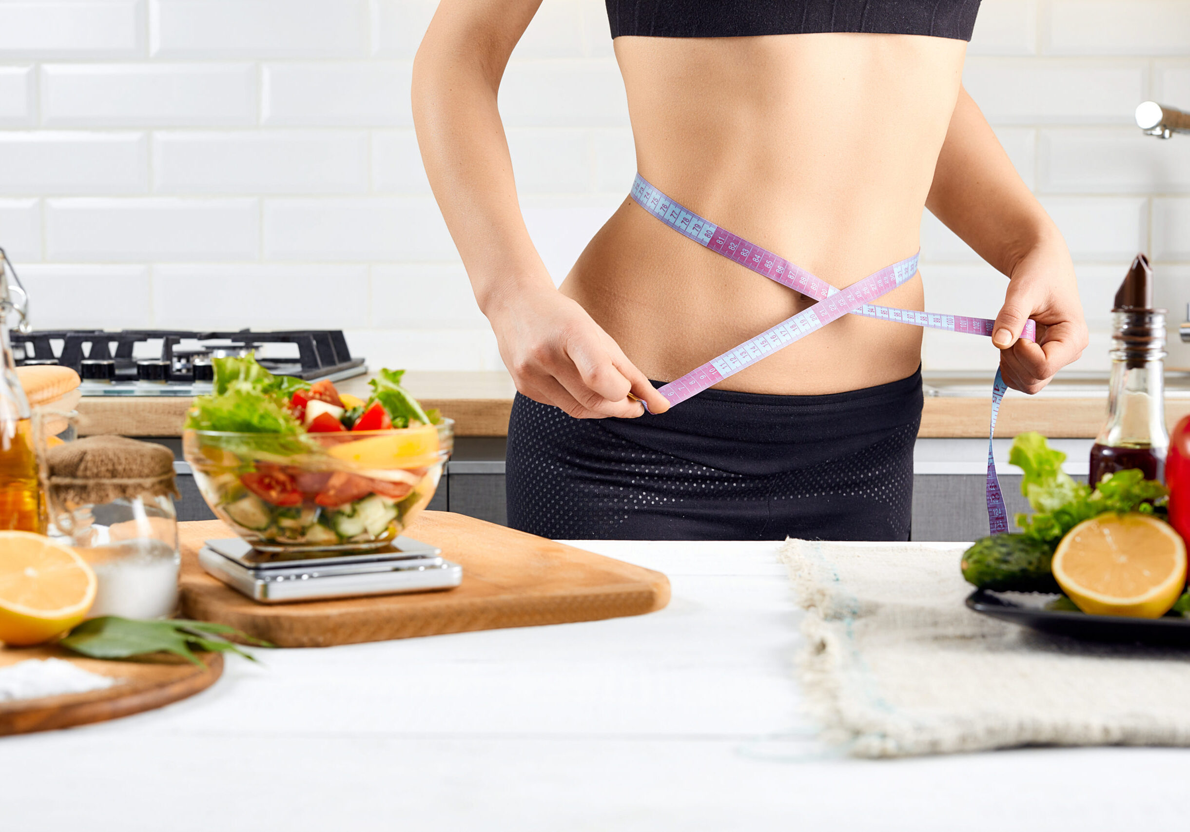 Diet, healthy eating, food and weigh loss concept. Young woman measuring waist near tomatoes, peppers and salad on the kitchen interior.